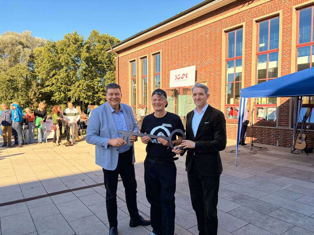 Links steht der Finanzsenator Andreas Dressel, in der Mitte ein Mensch vom Verein. Rechts steht ein weiterer Mann, der offiziell aussiehr. Im Hintergrund die sonnenbeschienene Backsteinfassade der Düse. Foto: Chris Meyer