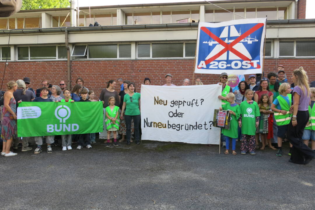 Viele Menschen vor einer Mauer halten Transparente. Von links ein grünes BUND-Banner, dann. ein weißes Transparent „Neu geprüft oder nur neu begründet" dann ein blau-weißer rot durchgekreuztes A26-Banner