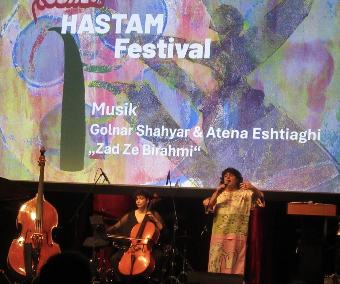 Links im Bild ein herrenloser Bass, in der Mitte sitzt Atena Eshtighi am Cello, rechts steht die Sängerin Golnasr Shahyar. Im Hintergrund das auf die Leinwand projizierte Festivalplakat
