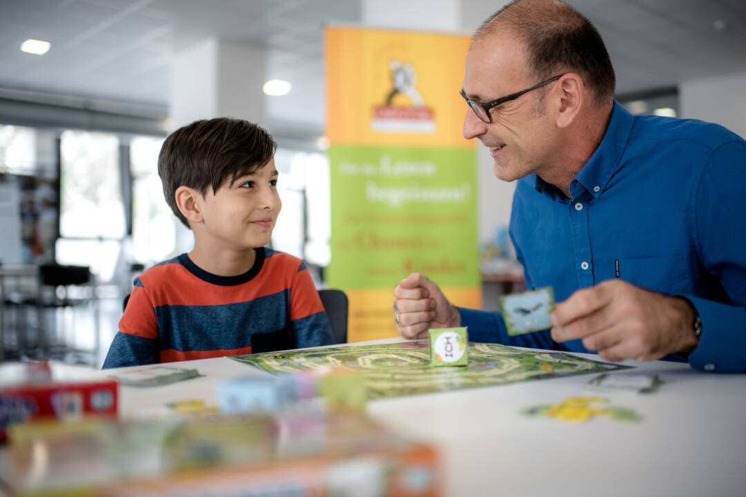 In einem großen, eher nüchternen Raum (Klassenzimmer?) sitzen an einem Tisch ein Kind und ein älterer Mann. Sie spielen. Es gibt einen besonderen Würfel mit Buchstaben und Kärtchen. Foto: Endermann