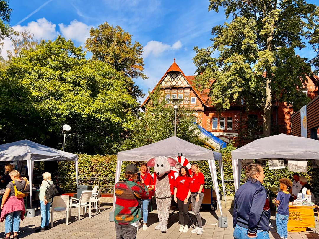 Vor einem Backsteinfachwerkhaus im Hintergrund und Bäumen und Hecken stehen auf einem Platz drei Pavillons, außerdem Stühle. Es sind mehrere Menschen im Bild. Im Zentrum steht eine Gruppe Haspa-Angestellte in roten Shirts um das Maskottchen der HASPA, eine Maus herum und wird fotografiert. Foto: Chris Meyer