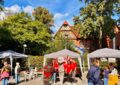 Vor einem Backsteinfachwerkhaus im Hintergrund und Bäumen und Hecken stehen auf einem Platz drei Pavillons, außerdem Stühle. Es sind mehrere Menschen im Bild. Im Zentrum steht eine Gruppe Haspa-Angestellte in roten Shirts um das Maskottchen der HASPA, eine Maus herum und wird fotografiert. Foto: Chris Meyer