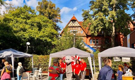 Vor einem Backsteinfachwerkhaus im Hintergrund und Bäumen und Hecken stehen auf einem Platz drei Pavillons, außerdem Stühle. Es sind mehrere Menschen im Bild. Im Zentrum steht eine Gruppe Haspa-Angestellte in roten Shirts um das Maskottchen der HASPA, eine Maus herum und wird fotografiert. Foto: Chris Meyer