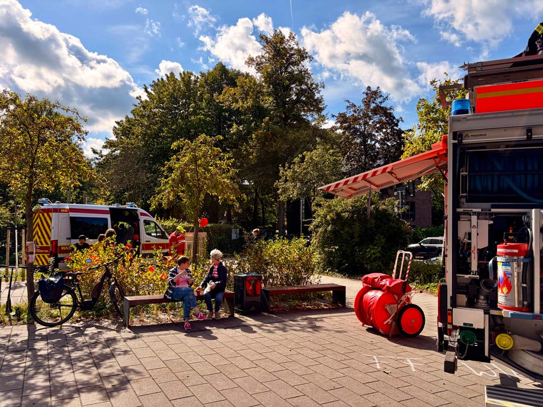 Man sieht gar nicht, dass es ein gut besuchtes Fest ist. Im Hintergrund ein Infostand der Deutschen LebensRettungsGesellschaft, im Vordergrund die hintere Ecke eines Feuerwehrautos. Dazwischen, auf einer Bank in der Sonne zwei Personen, die in ein angeregtes Gespräch vertieft sind. Foto: Chris Meyer