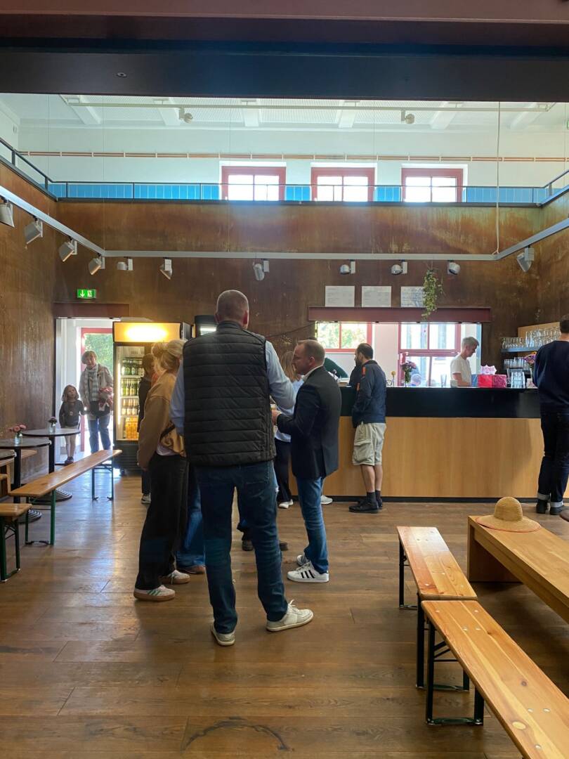 In einem braun getäfelten Raum auf Parkett stehen einige Menschen. Im Hintergrund eine Theke und rechts und links Biertischgarnituren.
