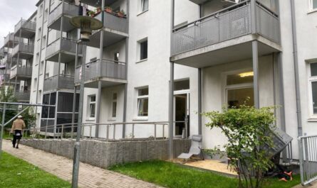 An einem Plattenweg stehen weiße Häuser mit angebauten metallenen Balkonen. Rechts unten eine kleine Standidylle mit Liegestuhl