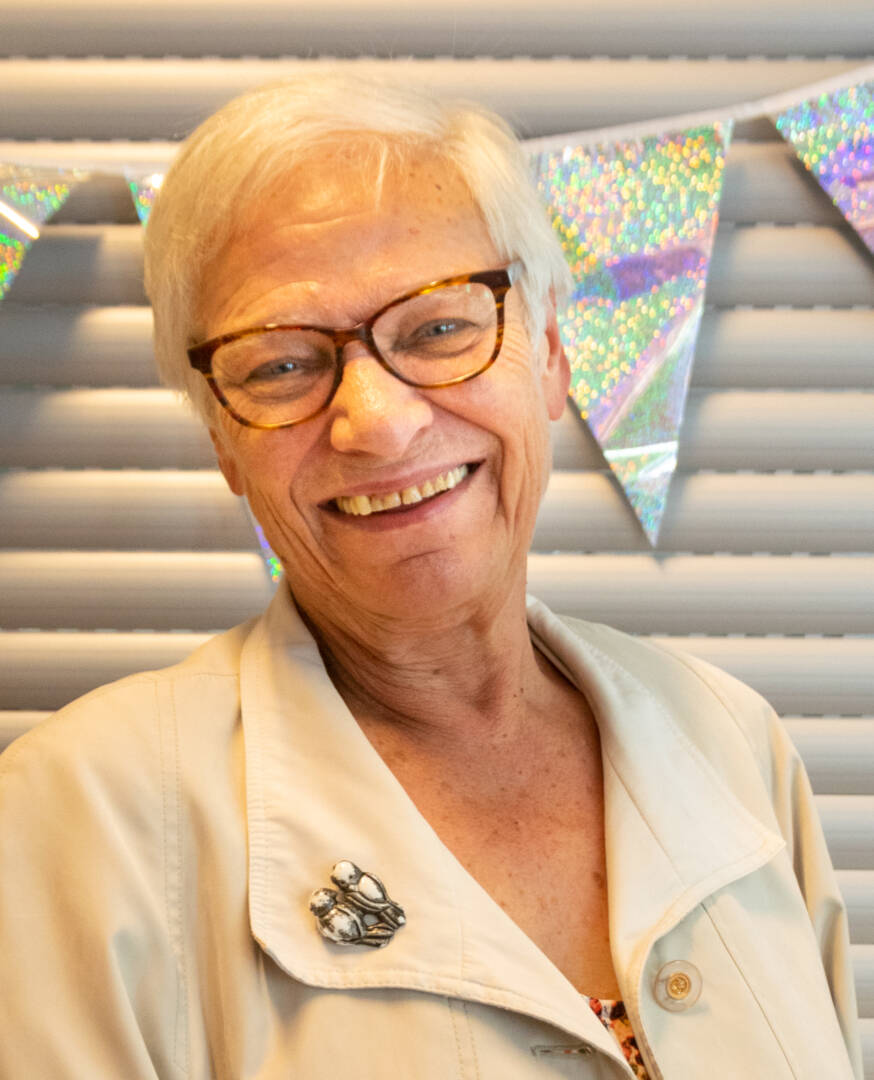 Portrait, Kopf und Schultern. Vor einer hellen Wand mit einer glitzernden Wimpelkette steht eine Frau. Sie hat sehr helle Haare, eine braune Brille und trägt eine sportlich-elegante Jacke mit einer großen Brosche. Sie strahlt in die Kamera.