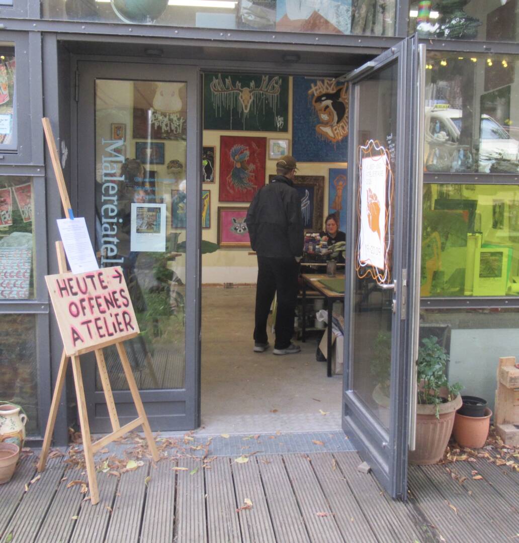 Der Eingang zum Atelier auf dem Hof der Honigfabrik, hinter einer Glastür erkennt amn unmengen von Bildern. Zwei Erwachsene im Gespräch.