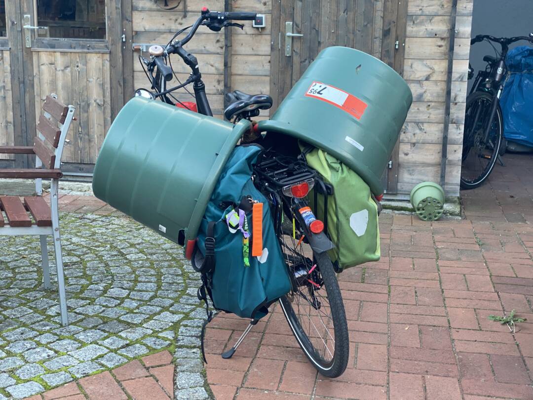 Auf einem gepflasterten Hof steht ein normales Zweirad. am Gepäckträger hängen Fahrradtaschen. Und über den Fahrradtaschen jeweils ein große grüne Gartentonne, vmtl. ca. 100 l fassend. In der Mitte sind die beiden Tonnen zuzusammengebunden. Wie sie am Rad befestigt sind (Spanngurt) ist nicht zu erkennen.