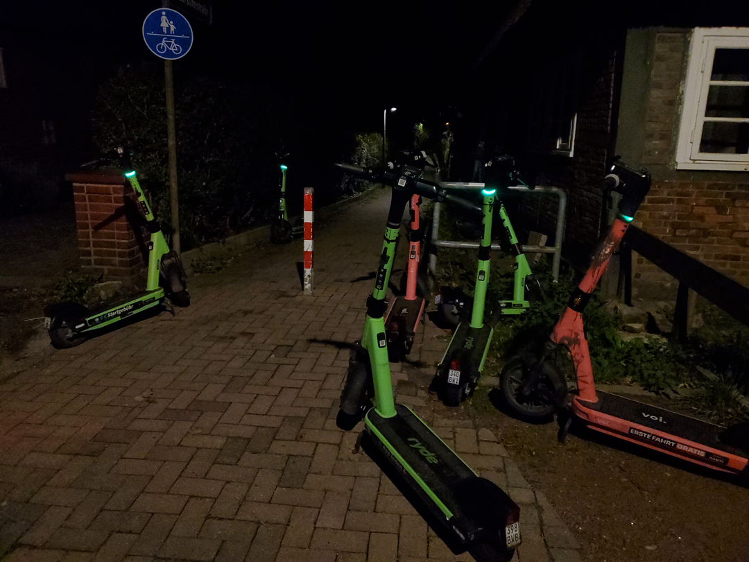 Nachtaufnahme: Auf einem schmalen Fußweg, der zusätzlich durch einen Poller verschmäler ist, stehen links und rechts insgesamt 7 E-Scooter. Rechts im Bild eines der ältesten Häuser in Wilhelmsburg, zu erkennen ist eine Fachwerk-Backsteinwand und ein weißer Fensterrahmen
