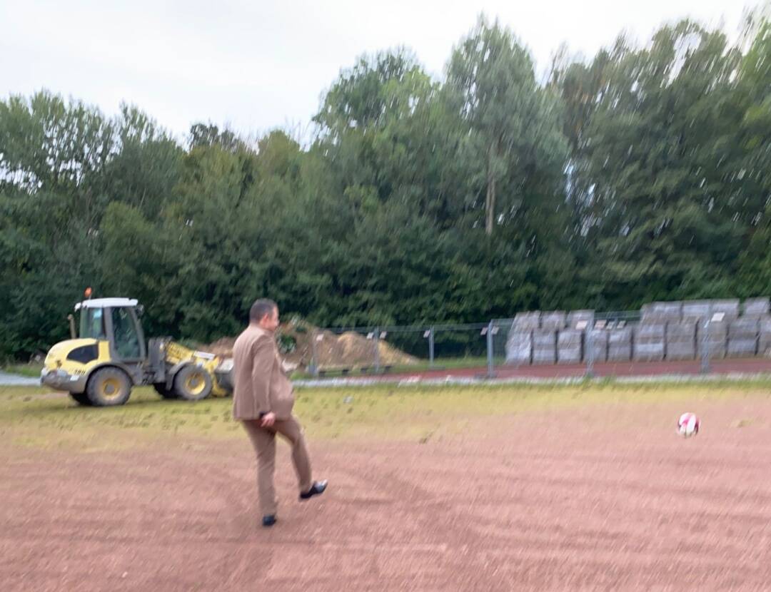 In Hintergrund ein Bagger und Paletten mit Pflastersteinen. Im Vordergrund ein Mann und rechts davon ein Ball