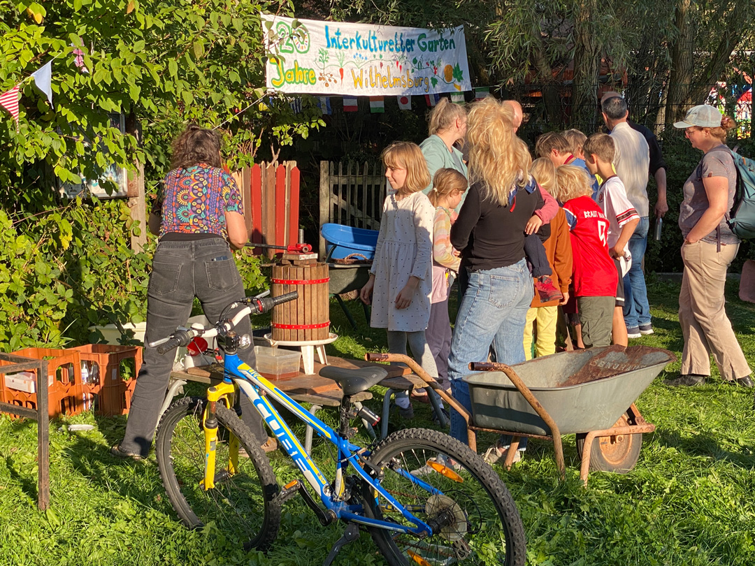Auf dem Rasen vor dem Interkulturellen Garten sind Kinder und Erwachsene dabei, von Hand Apfelsaft herzustellen: Waschen, schneiden, häckseln, pressen. Drum herum ein Fahrrad, eine Schubkarre, leere Flaschen.