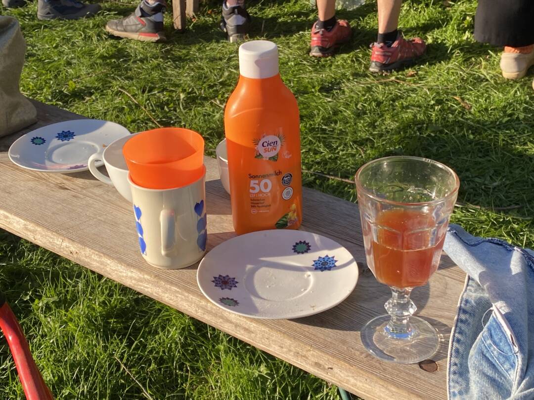 Im Frühherbst. Auf einer Holzbank steht ein lustiges Stilleben: Becher Teller, ein Glas mit orangefarbenem Apfelsaft und Sonnenmilch. Die Sonnenmilchflasche und ein Becher sind ähnlich orange wie der Apfelsaft.