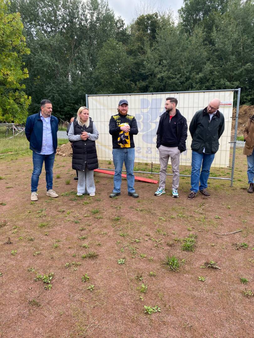 Vor einer weißen Plastikplane stehen fünf Menschen auf einem Tennenplatz
