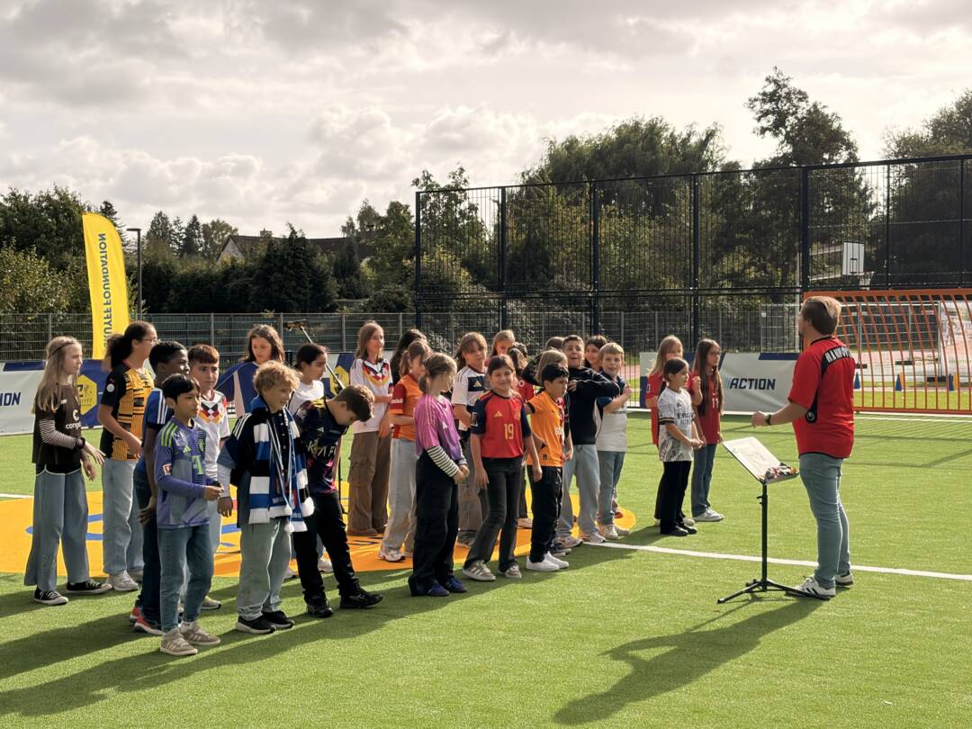 Eine Gruppe Kinder auf dem neuen sattgrünen Kunstrasenplatz. Rechts steht ein Erwachsener. Entweder dirigiert er oder er hält eine Rede.