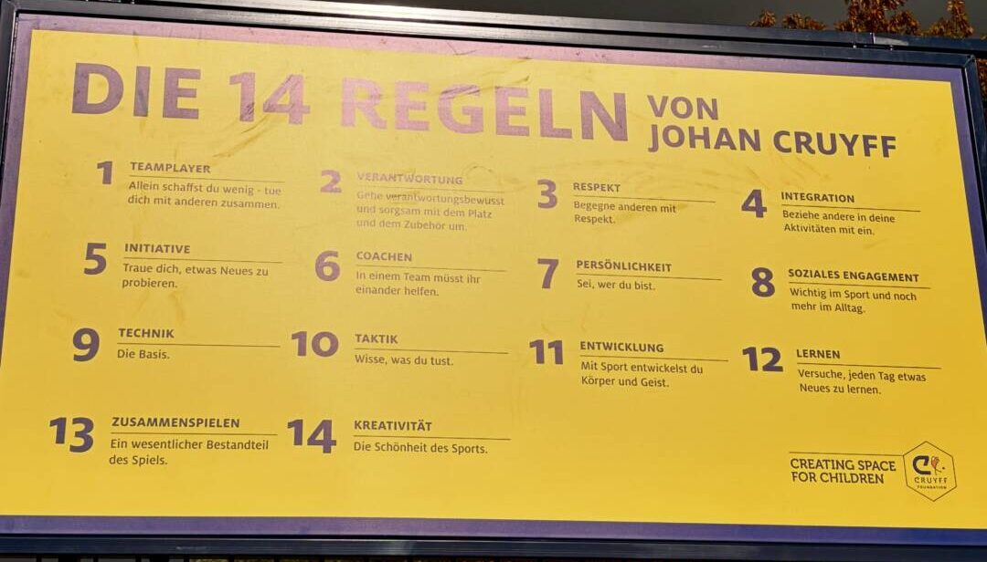 Gelbes Schild mit den Regeln von Johan Cruyff: erstens Team Player, zweitens Verantwortung, drittens Respekt, viertens Integration, fünftens Initiative, sechstens Coaching, 7. Persönlichkeit, achtens soziales Engagement, neuntens Technik, zehnten Taktik, elftens Entwicklung, zwölftens lernen, 13 tens Zusammenspielen, 14. Kreativität