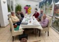 Eine Frau und vier Männer sitzen um einen gedeckten Tisch mit weißer Tischdecke. Hinten an der Wand ist eine große Luftballon-76.