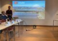 Ein Mann steht links hinter einem Tisch neben dem Eröffnungsbild der Präsentation
