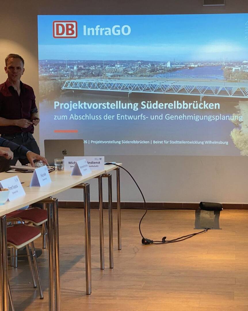 Links steht ein Mann hinter einem Tisch. Rechts daneben die visuelle Präsentation der Projektvorstellung.