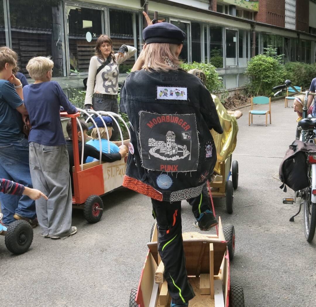 8 Kinder, 2 Erwachsene, 3 unterschiedliche Holzfahrzeuge und ein Fahrrad vor einem Gebäude, das aussieht wie eine Schule aus den 1970ern.