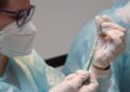 Ein Mensch mit weißer Maske und dunkler Brille zieht eine Spiritze auf. Er/Sie trägt einen Plastikschutz über einem weißen Kittel und Gummihandschule.