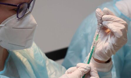 Ein Mensch mit weißer Maske und dunkler Brille zieht eine Spiritze auf. Er/Sie trägt einen Plastikschutz über einem weißen Kittel und Gummihandschule.