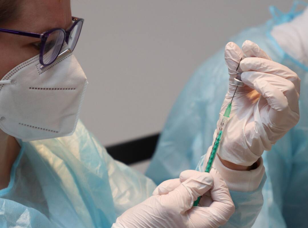 Ein Mensch mit weißer Maske und dunkler Brille zieht eine Spiritze auf.
Er/Sie trägt einen Plastikschutz über einem weißen Kittel und Gummihandschule.